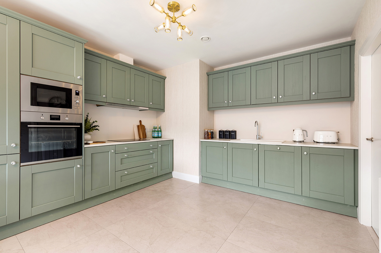 Modern kitchen with green cabinetry, built-in oven and microwave, white countertops, and minimalist decor including toaster, kettle, and jars.