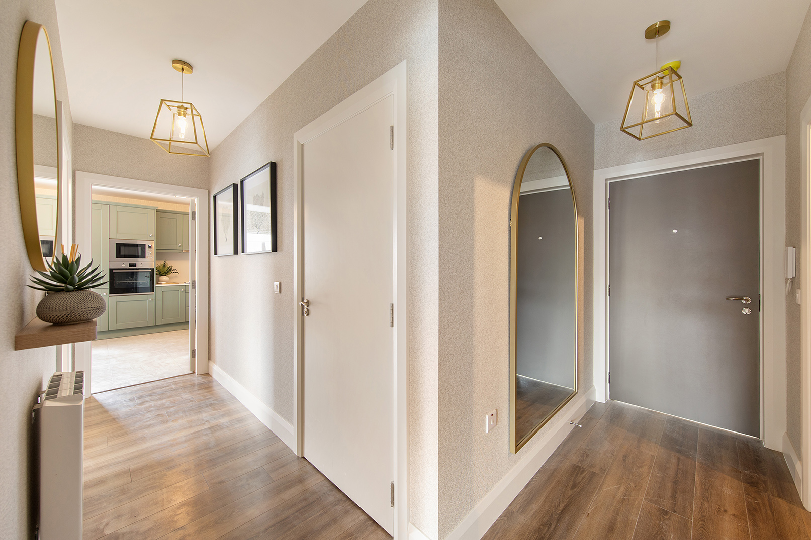 Modern hallway with wood flooring, gray front door, large arched mirror, gold pendant light, and a view into a kitchen with sage green cabinets.