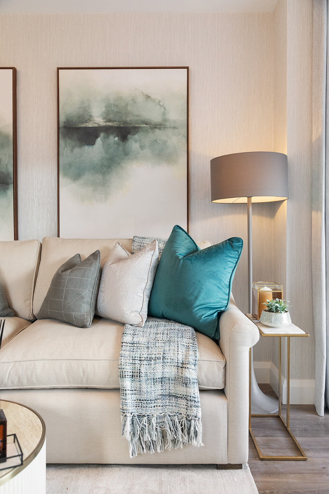 Beige sofa with teal, gray, and light pink cushions and a blue-gray throw blanket, next to a gold and white side table with a lamp and a small plant in a modern living room.