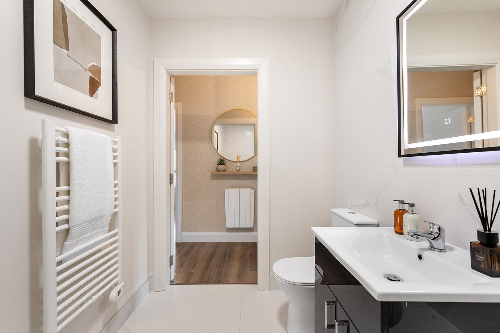 Modern bathroom with white walls, a towel radiator with a white towel, a toilet, a dark vanity with a white sink, and a mirror reflecting the doorway.